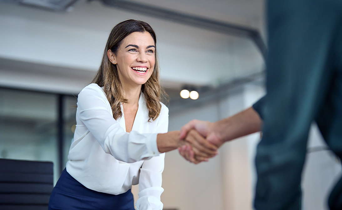 Leende kvinna i affärsmiljö skakar hand med person.